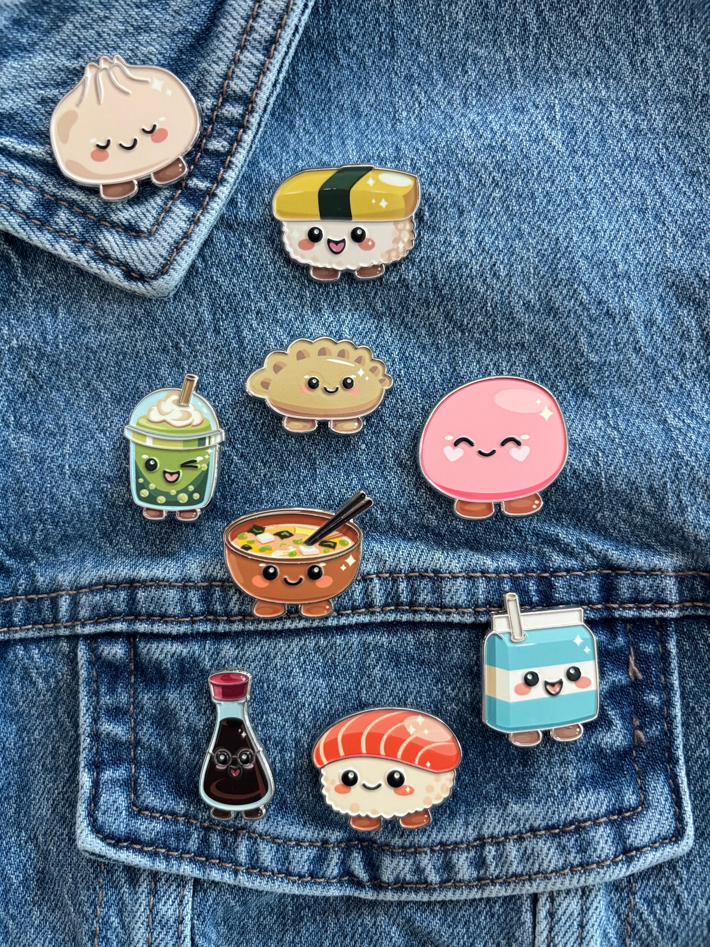 Nine cute sushi-themed enamel pins are arranged on a blue denim jacket pocket. The pins feature cartoon characters resembling a kind dumpling, sunny egg maki wrap, bubbly matcha tea, gentle mochi, patient bowl of ramen, smart soy sauce bottle, sweet salmon sushi, cheerful gyoza and a cool milk carton.
Copyright © Cute Bites® All rights reserved.