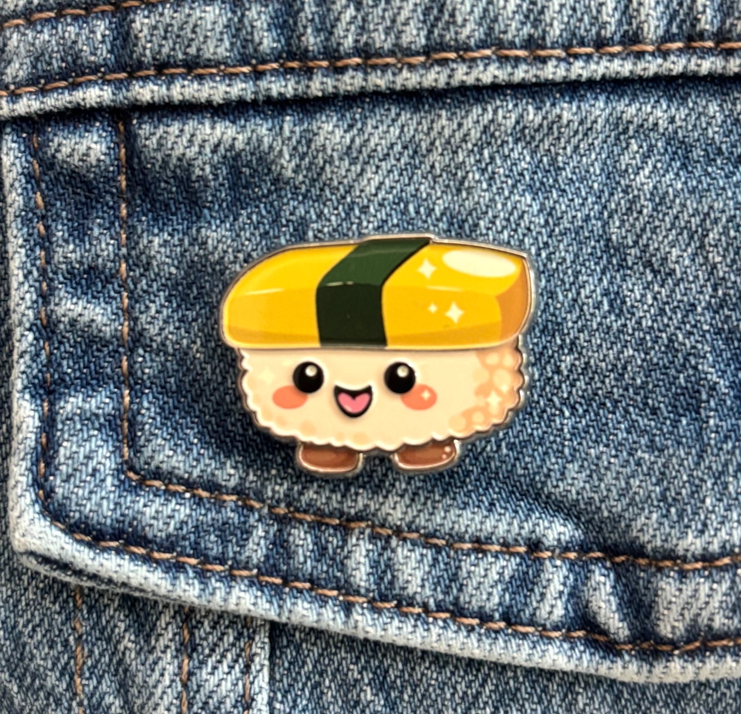 A cute kawaii sunny egg maki wrap sushi-shaped enamel pin with a smiling face, placed on a blue denim pocket. Copyright © Cute Bites® All rights reserved.