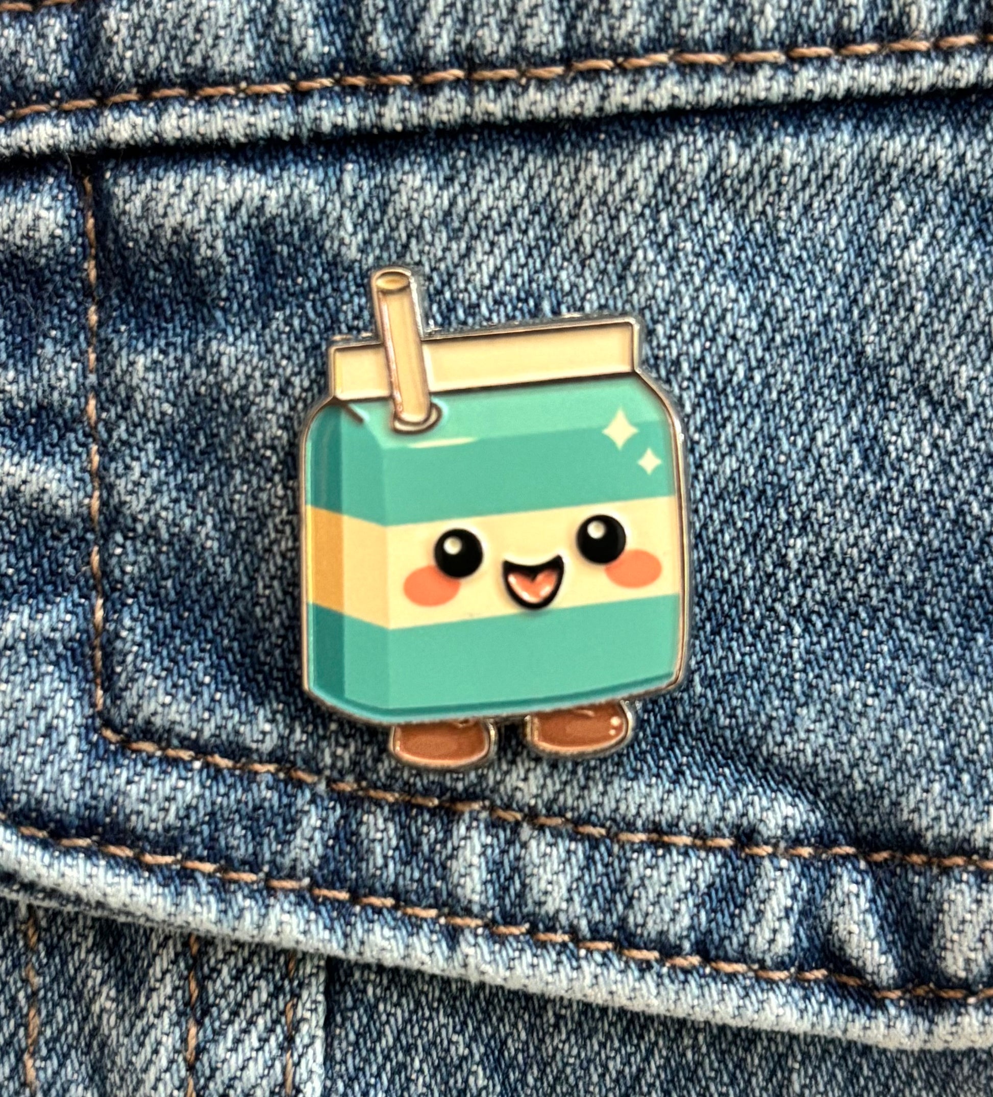 A cute kawaii-style enamel pin shaped like a smiling cool milk carton with a straw, shown pinned onto blue denim fabric.
Copyright © Cute Bites® All rights reserved.