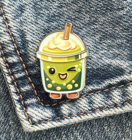A cute enamel pin shaped like a winking green bubble matcha drink cup with whipped cream and a straw, placed on blue denim fabric.