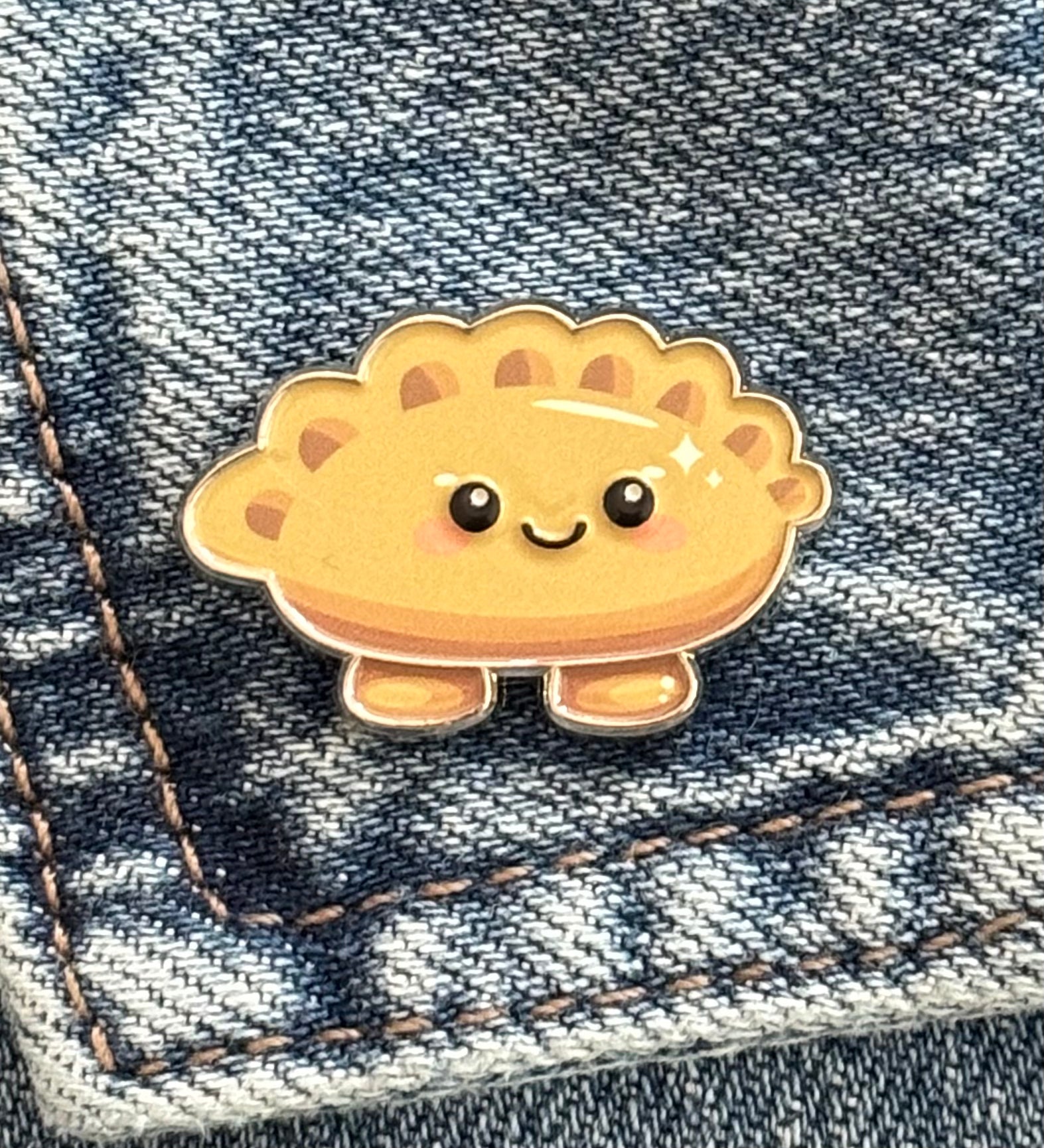 A cute, cartoon-style golden-brown cheerful gyoza pin with a smiling face, shown pinned onto a blue denim jacket.
Copyright © Cute Bites® All rights reserved.